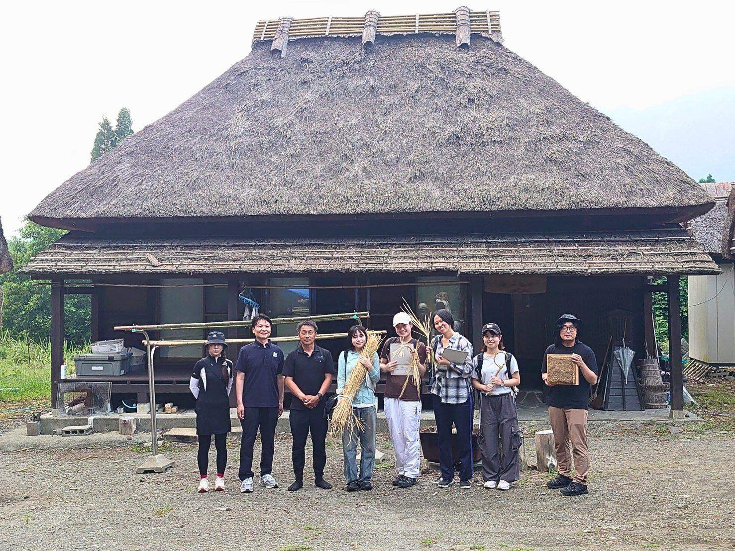 阿蘇茅葺工房の茅葺き屋根の民家の前で、茅の束を手にした学生たちと一行の集合写真