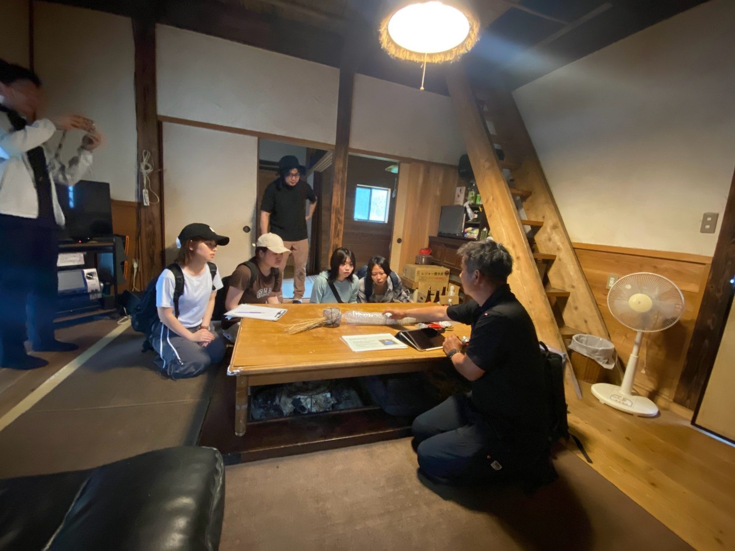 茅葺き民家の囲炉裏のある座敷で、職人さんの話を聞く学生たち