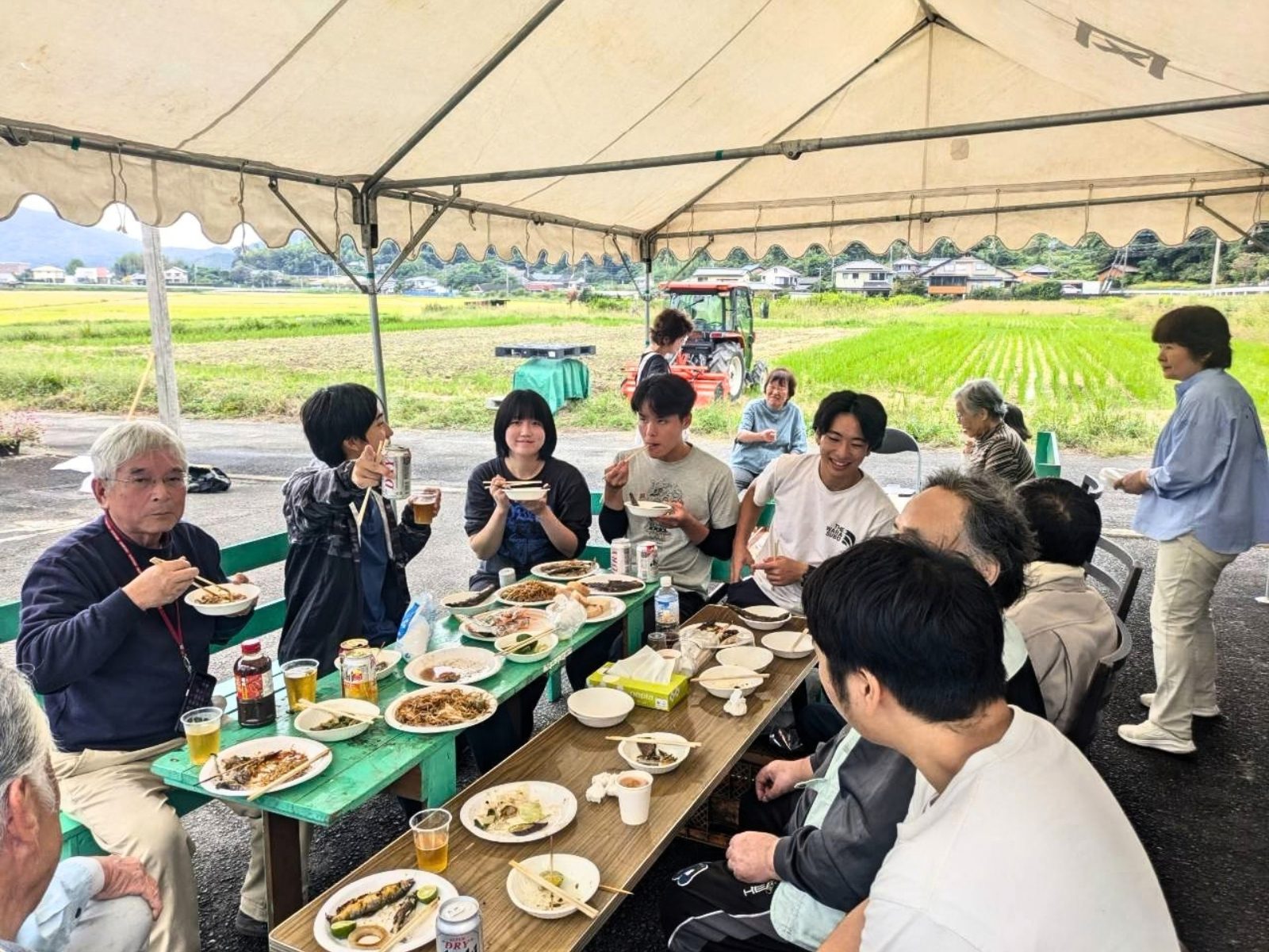 田んぼを望むテントの下で、地域のみなさんと学生が長テーブルを囲んで鮎や料理を食べている