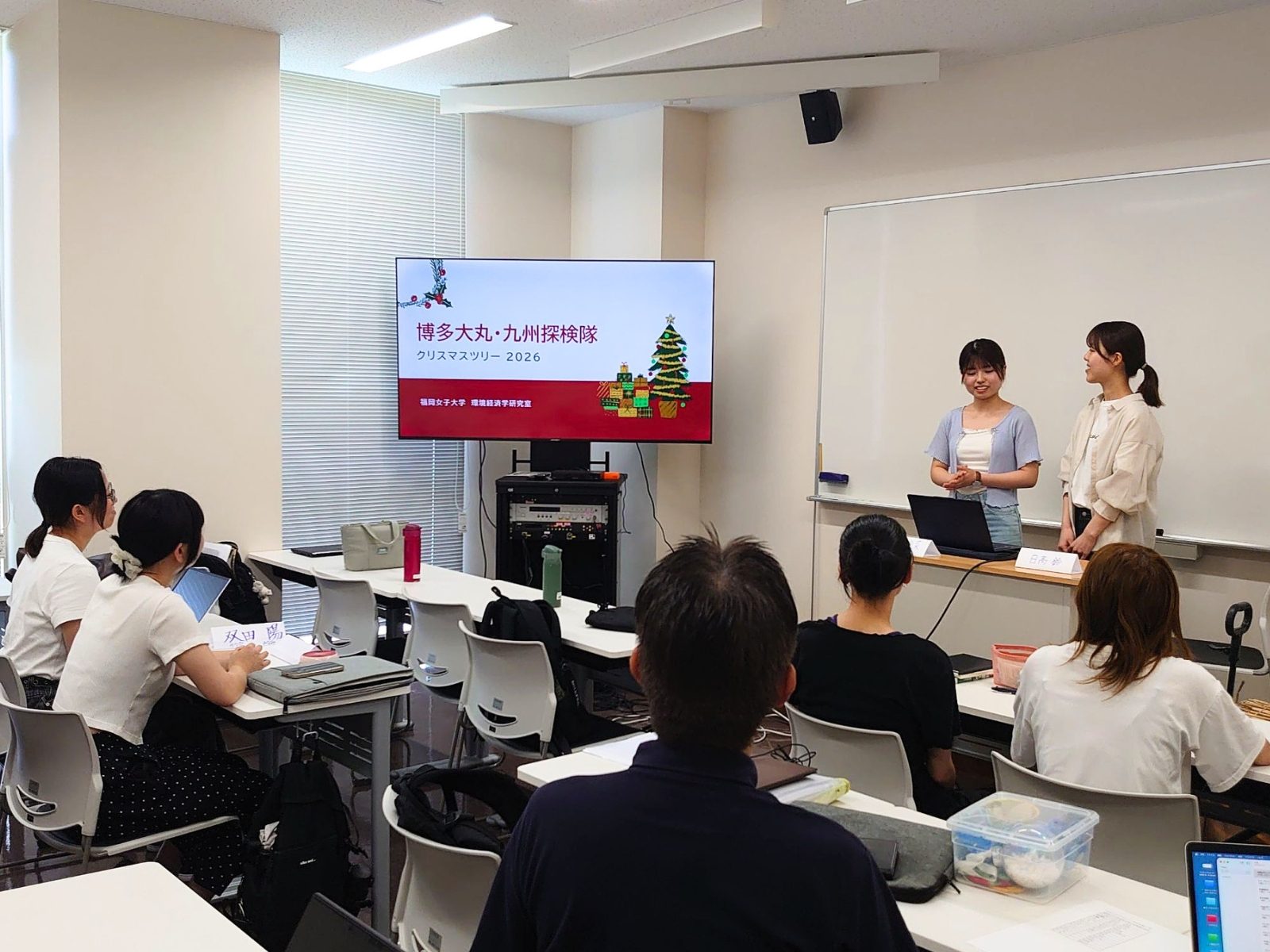 福岡女子大学の教室で「博多大丸・九州探検隊 クリスマスツリー2026」のタイトルスライドを背に発表する学生2人と、それを聞く参加者