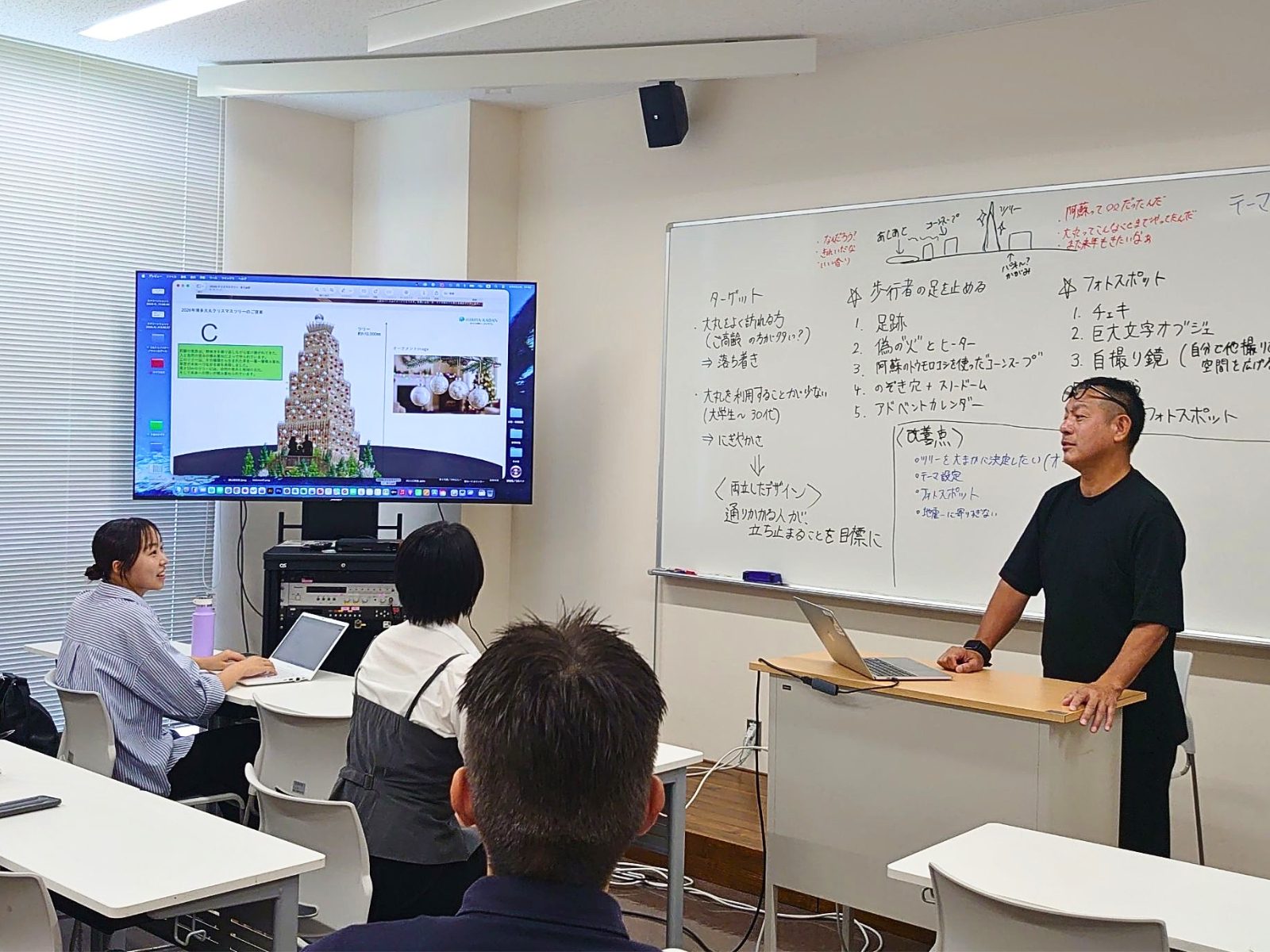 福岡女子大学の教室で、ツリーのデザイン案を映したモニターとホワイトボードを前に話す大丸福岡天神店のメンバーと、それを聞く学生たち