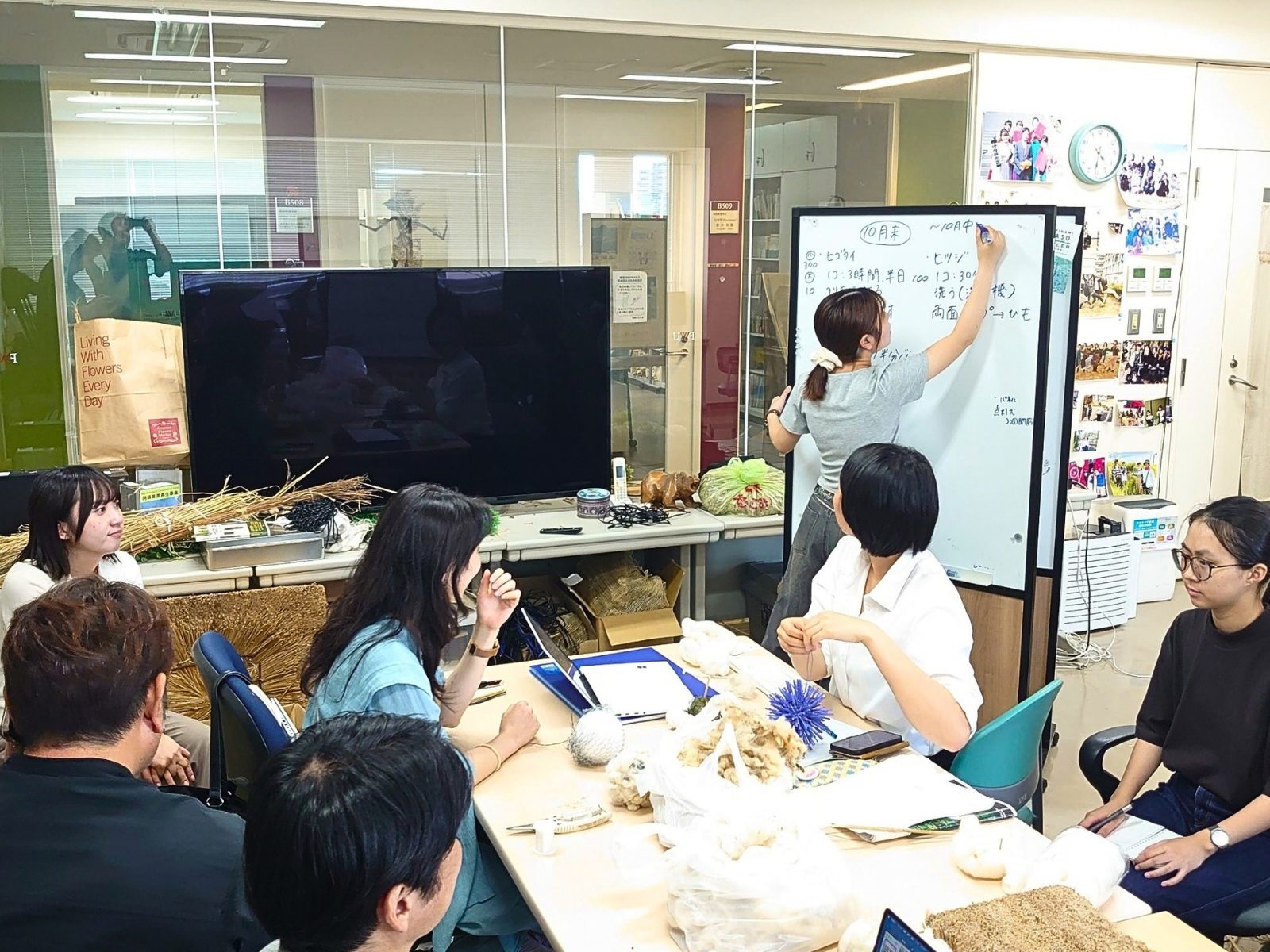 福岡女子大学の研究室で、茅や羊毛などの素材と試作品を机に広げ、ホワイトボードに書き込みながら話し合う学生と大丸福岡天神店・唐津Farm&Foodのメンバー