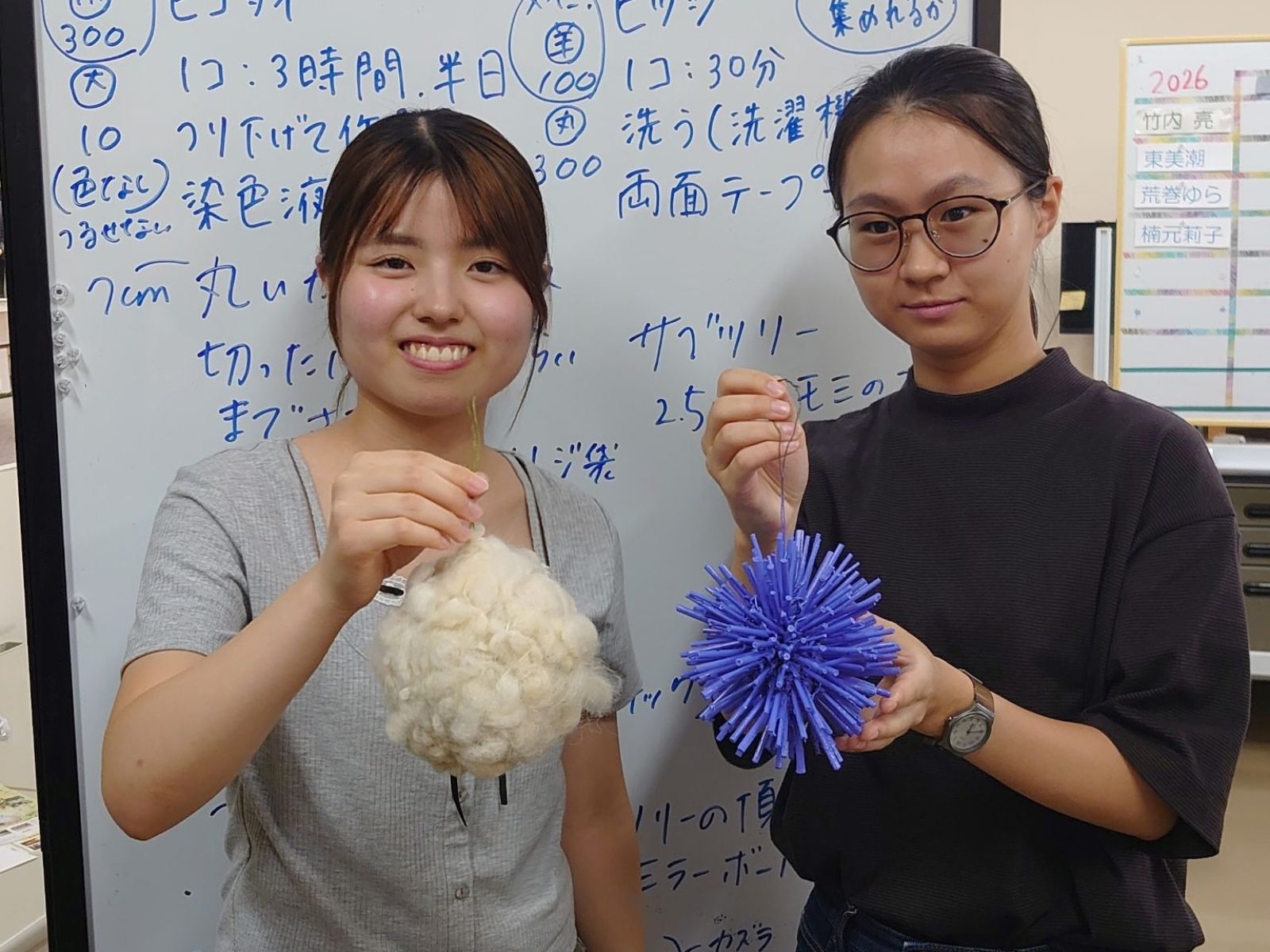 製作の段取りを書き込んだホワイトボードの前で、羊毛のオーナメントと紫色の試作オーナメントを手に持つ福岡女子大学の学生2名