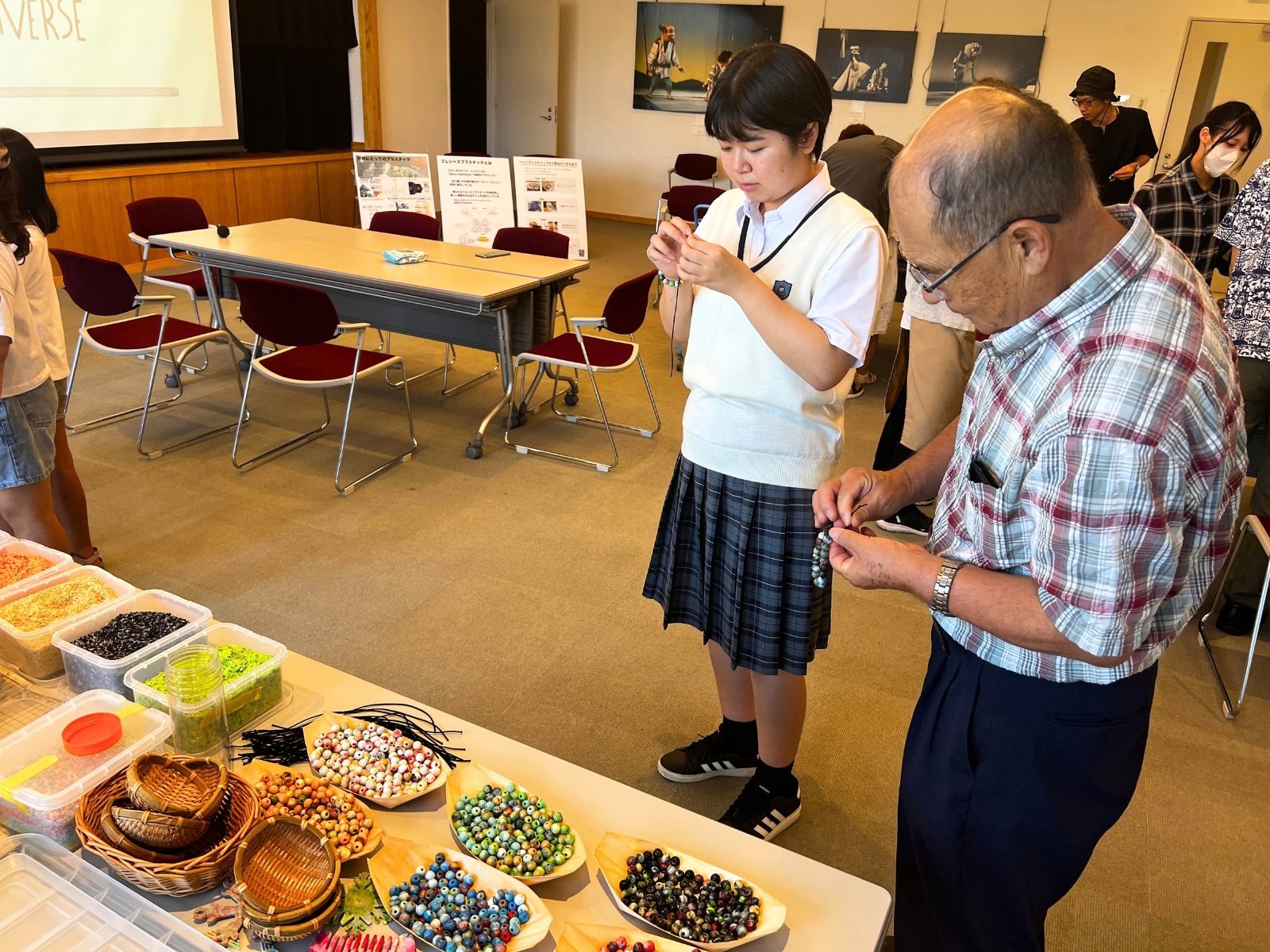 色とりどりのリサイクルプラスチックビーズを並べたテーブルの前で、ブレスレットづくりに取り組む学生と年配の参加者