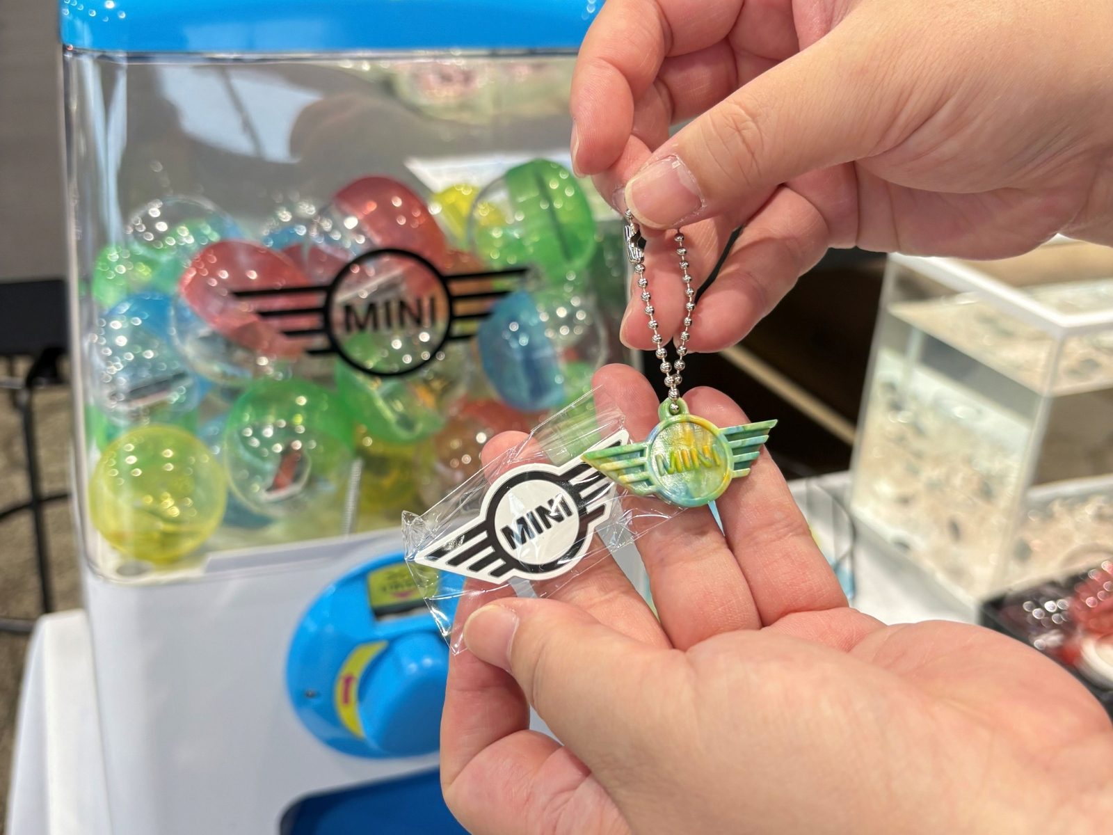 カプセルが詰まったMINIロゴ入りのガチャガチャの前で、手のひらにのせたMINIロゴキーホルダーとステッカー