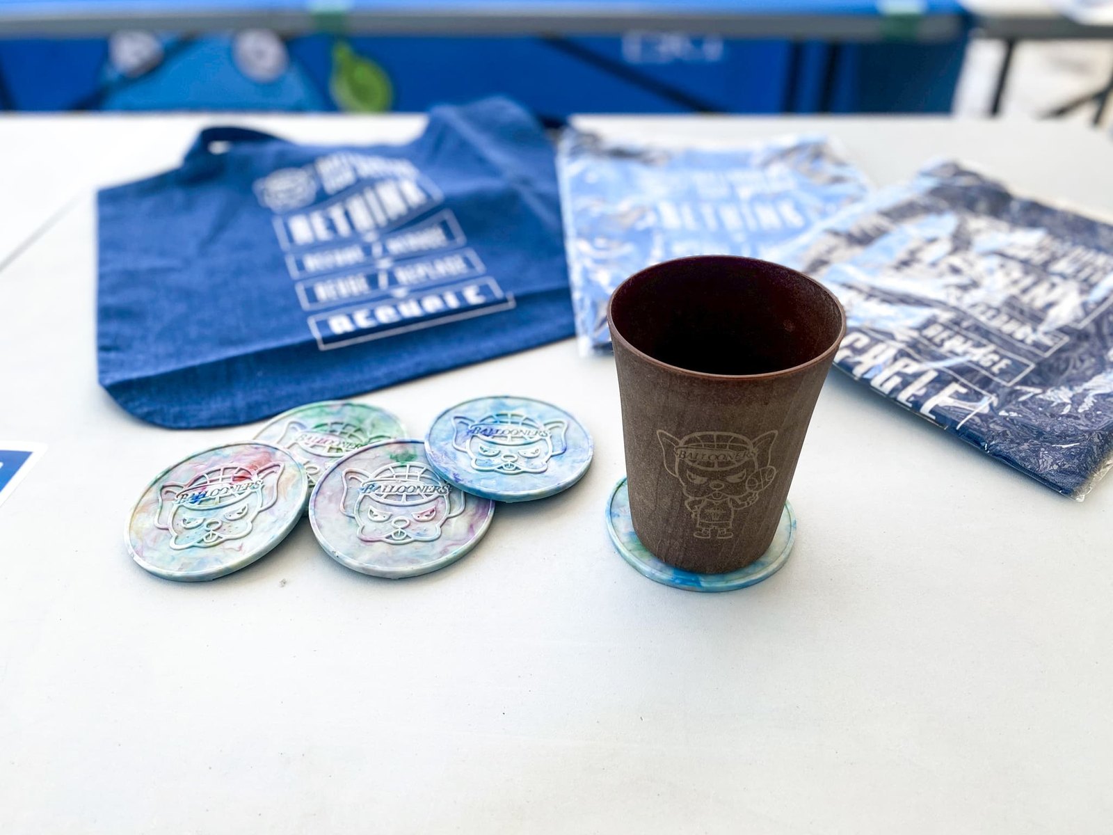 A coaster printed with the Baltan mascot, with a tote bag and a cup