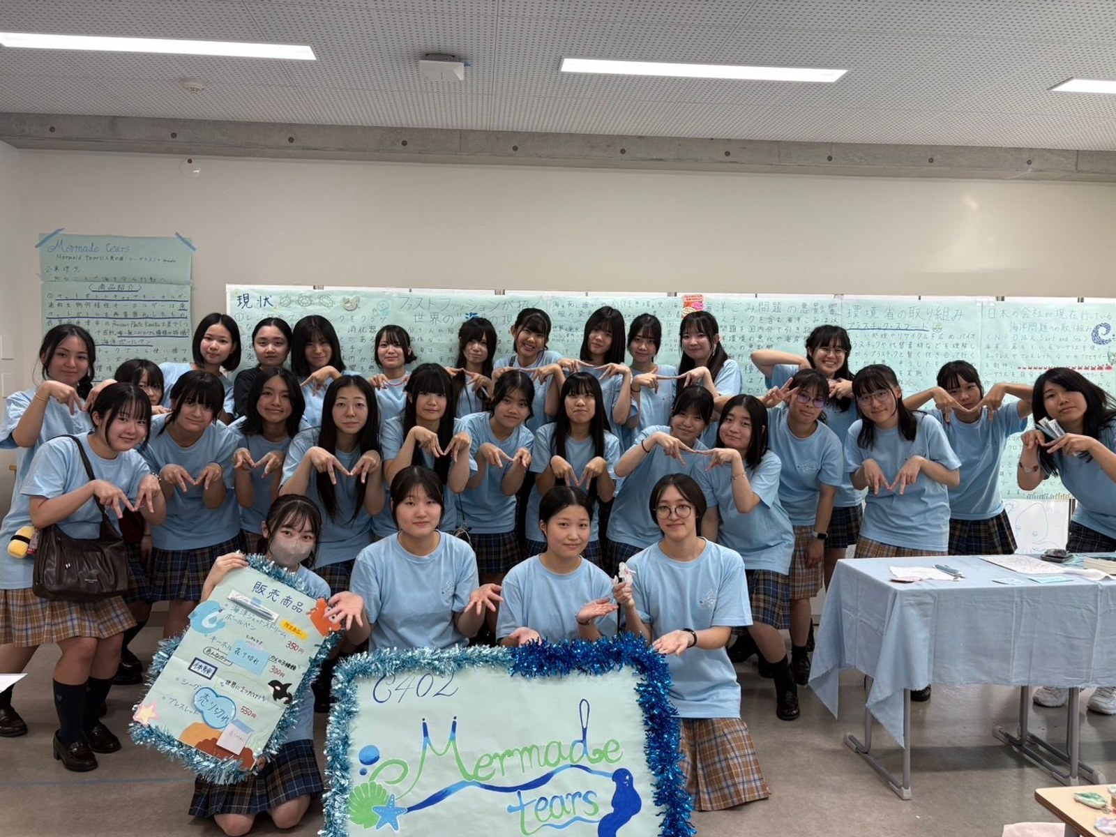 「Mermade tears」の看板と販売商品のボードを持ち、そろいの水色のTシャツで集合した品川女子学院の生徒のみなさん