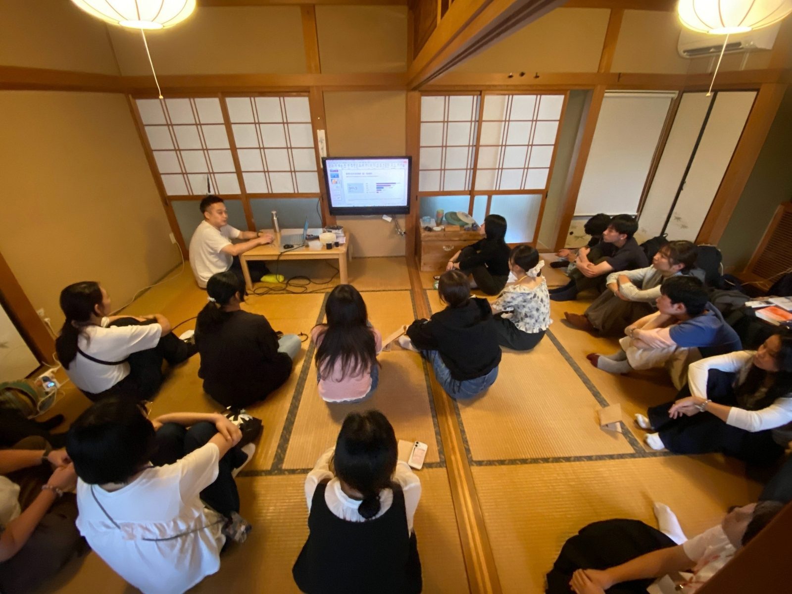畳の部屋で、スライドを示しながら話す講師と、それを囲んで聞く参加者たち