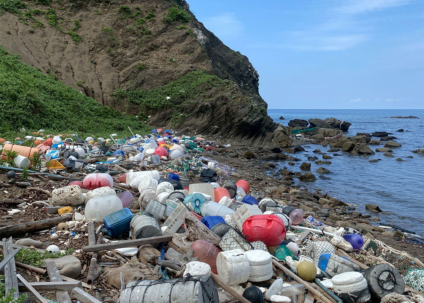 A Tsushima cove buried in drifted marine debris, with steep forested cliffs behind